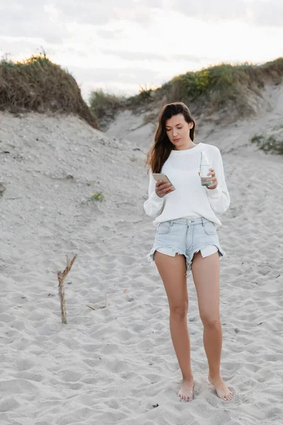 Barefoot woman using smartphone and holding bottle of water on beach — Stock Photo