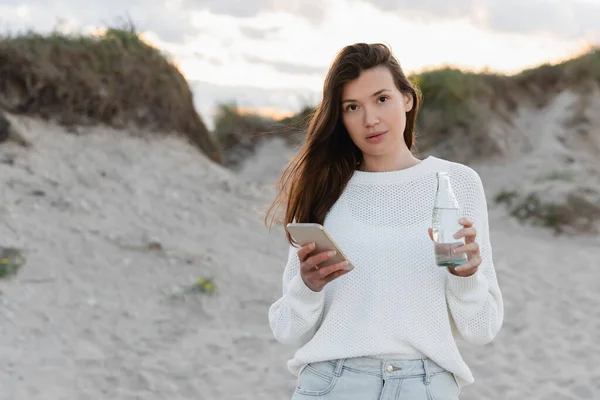 Brunette woman holding smartphone and bottle of water on beach in evening — Stock Photo