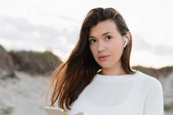 Bruna donna in auricolare in possesso di smartphone e guardando la fotocamera sulla spiaggia — Foto stock