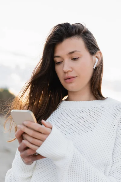 Brunette woman in wireless earphone using blurred smartphone on beach — Stock Photo