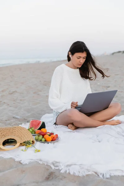 Brunette woman in earphone using laptop near fruits on blanket — Stock Photo