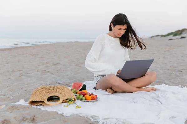 Freelance dans l'écouteur à l'aide d'un ordinateur portable près des fruits et sac à main sur la plage le soir — Photo de stock