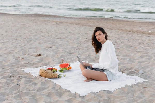 Joven freelancer en auriculares con portátil cerca del bolso y frutas frescas en la playa - foto de stock