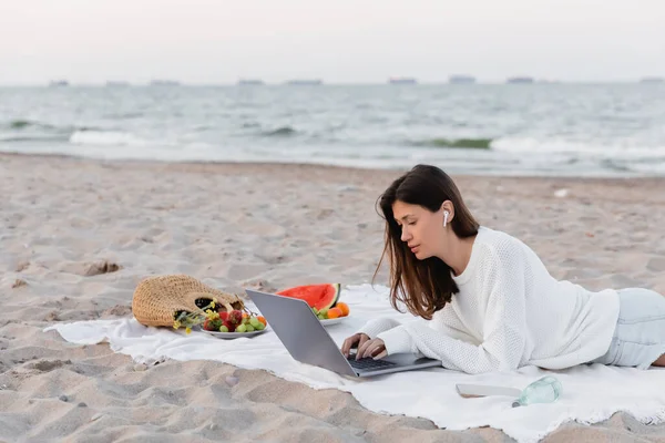 Woman in earphone using laptop near smartphone and fruits on blanket on beach — Stock Photo