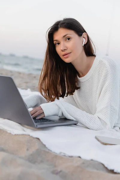 Freelancer in earphone using laptop and looking at camera near smartphone on blanket on beach — Stock Photo