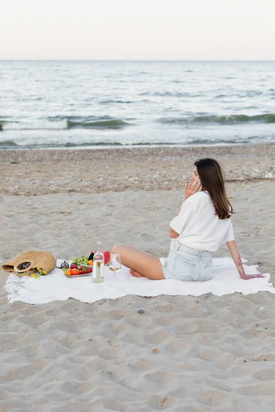 Mujer hablando en smartphone cerca de vino y frutas en la costa del mar - foto de stock
