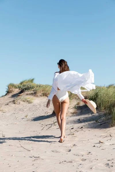 Rückansicht einer Frau in Badeanzug und weißem Hemd, die mit Strohhut am Sandstrand spaziert — Stockfoto