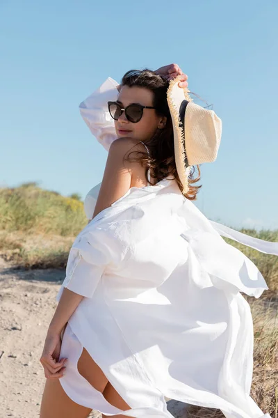 Mujer joven en gafas de sol y camisa blanca sosteniendo sombrero de paja en la playa de arena - foto de stock