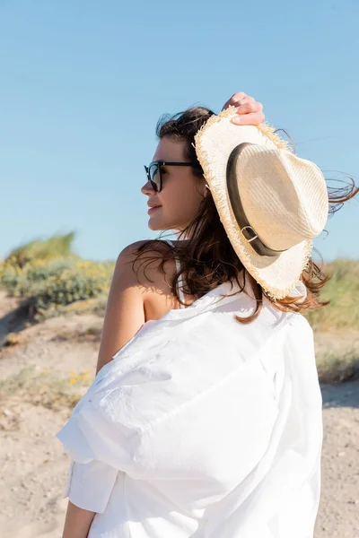 Vista lateral de mujer joven feliz en gafas de sol y camisa blanca sosteniendo sombrero de paja en la playa de arena - foto de stock