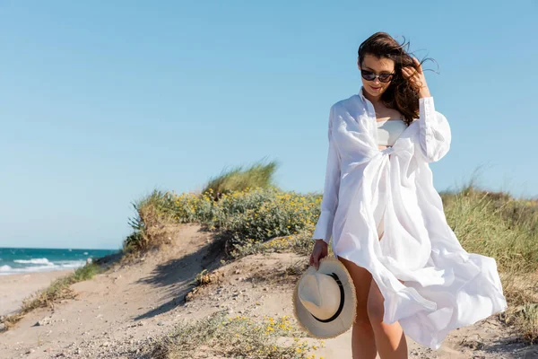 Mujer joven sonriente en gafas de sol y camisa blanca sosteniendo sombrero de paja en la playa de arena - foto de stock
