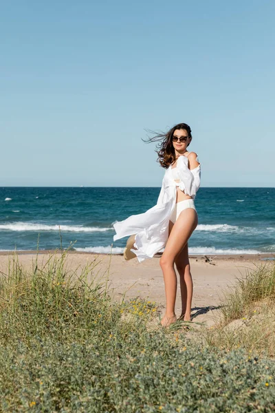 Pleine longueur de brune jeune femme en lunettes de soleil et chemise blanche tenant chapeau de paille sur la plage de sable — Photo de stock