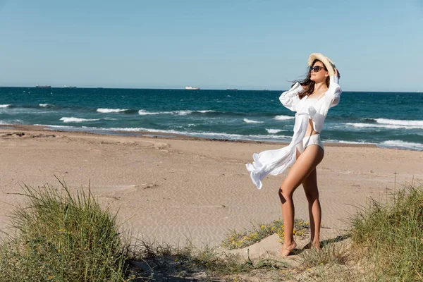 Полная длина стильная молодая женщина в солнечных очках и белой рубашке держа соломенную шляпу на песчаном пляже — стоковое фото