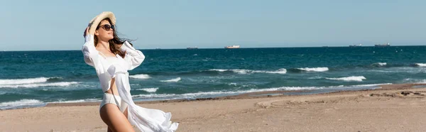 Усміхнена жінка в сонцезахисних окулярах і солом'яному капелюсі, що стоїть у білій сорочці біля моря на пляжі, банер — стокове фото