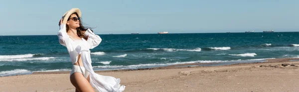 Smiling woman in sunglasses and straw hat standing in white shirt near sea on beach, banner — Stock Photo