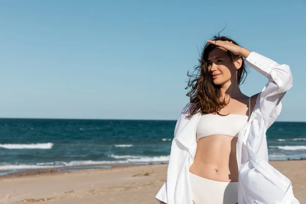 Giovane donna positiva in camicia bianca e costume da bagno vicino al mare blu sulla spiaggia di sabbia — Foto stock