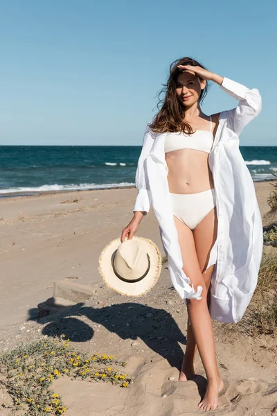 Sonriente mujer joven en camisa blanca sosteniendo sombrero de sol en la playa cerca del mar azul - foto de stock