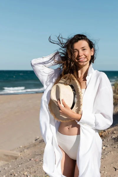 Cheerful young woman in white shirt and swimsuit holding straw hat on beach near sea — Stock Photo