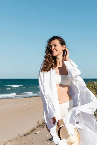 Giovane donna sorridente in camicia bianca e costume da bagno con cappello di paglia sulla spiaggia vicino al mare — Foto stock