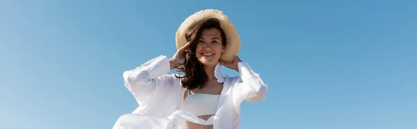 Vista basso angolo di felice giovane donna in costume da bagno e camicia bianca indossando cappello di paglia contro il cielo blu, banner — Foto stock