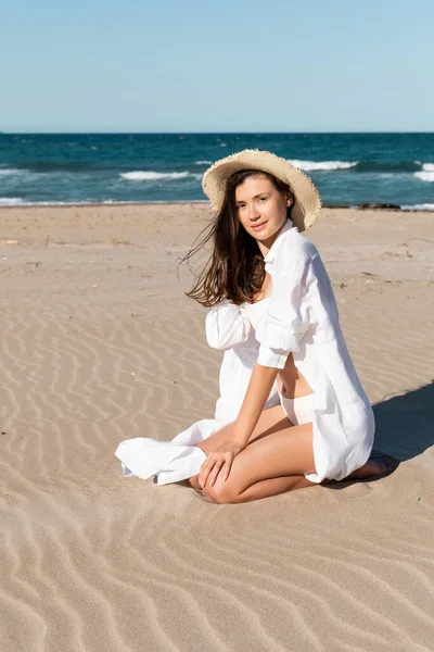 Jovem morena de chapéu de palha sentado na areia dourada perto do oceano azul — Fotografia de Stock