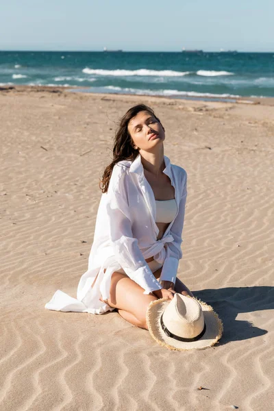 Jovem mulher de camisa branca e roupa de banho segurando chapéu de palha e sentado na areia dourada perto do oceano azul — Fotografia de Stock