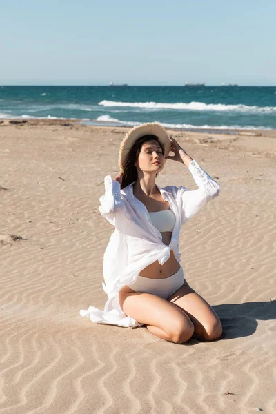 Young woman in white shirt and swimwear wearing straw hat and sitting on golden sand near blue ocean — Stock Photo