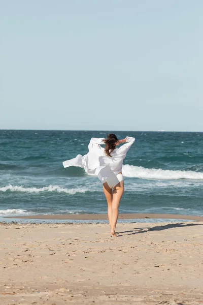 Vista trasera de la mujer joven en camisa blanca y traje de baño de pie cerca del océano en la playa - foto de stock
