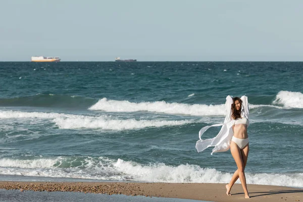 Longitud completa de la mujer joven en camisa blanca y trajes de baño de pie cerca del océano en la playa - foto de stock