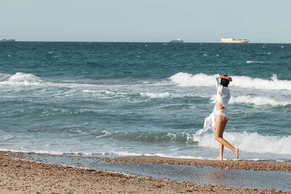 Volle Länge der jungen Frau in weißem Hemd und Badebekleidung, die am Strand am Meer spazieren geht — Stockfoto