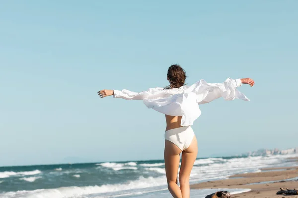 Rückansicht einer jungen Frau in weißem Hemd und Badebekleidung, die mit ausgestreckten Händen in der Nähe des Ozeans am Strand steht — Stockfoto