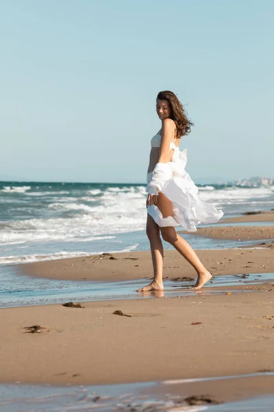 Piena lunghezza della donna a piedi nudi in camicia bianca e costumi da bagno in piedi vicino all'oceano sulla sabbia bagnata — Foto stock