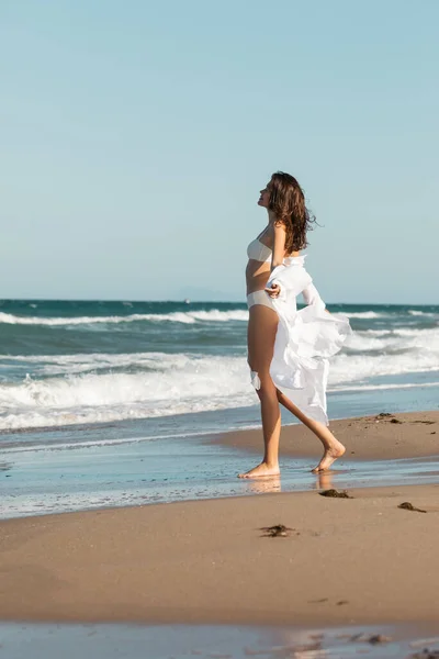 In voller Länge glückliche Frau in weißem Hemd und Badebekleidung, die am Meer auf nassem Sand steht — Stockfoto