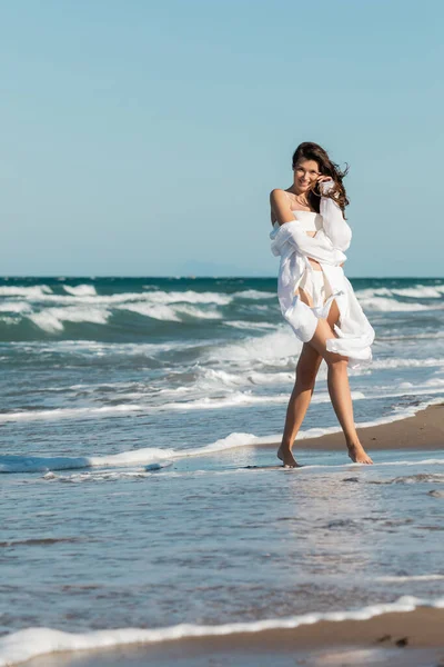 Comprimento total de mulher alegre em camisa branca e roupa de banho em pé perto do oceano na praia — Fotografia de Stock