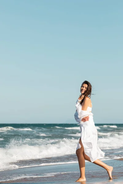 Piena lunghezza di donna gioiosa in camicia bianca e costume da bagno sorridente vicino oceano sulla spiaggia — Foto stock