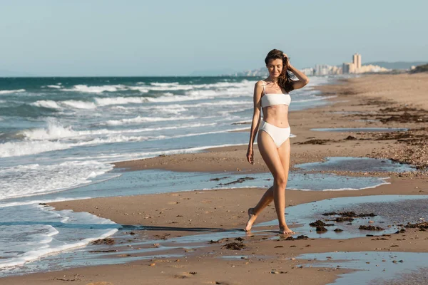 Longitud completa de la mujer alegre en traje de baño blanco caminando cerca del océano en la playa - foto de stock