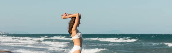 Vista lateral de la mujer feliz en traje de baño blanco caminando cerca del océano, bandera - foto de stock