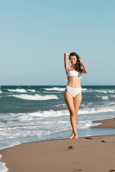 Longueur pleine de sourire jeune femme en maillot de bain blanc marchant près de l'océan sur la plage — Photo de stock