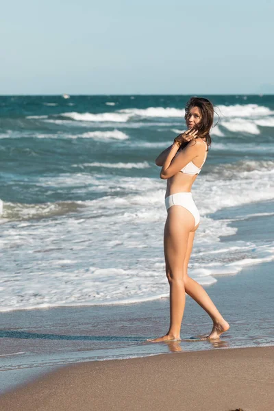 Longitud completa de morena joven en traje de baño blanco caminando cerca del océano en la playa - foto de stock