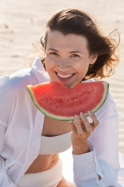 Retrato de mujer joven alegre en camisa blanca sosteniendo sabrosa sandía en la playa de arena - foto de stock