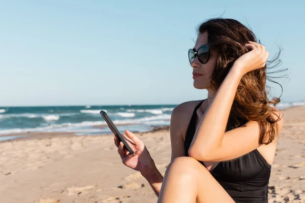 Jovem mulher em maiô e óculos de sol segurando smartphone perto do mar — Fotografia de Stock
