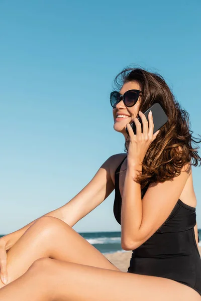 Happy young woman in swimsuit and sunglasses talking on smartphone near sea — Stock Photo