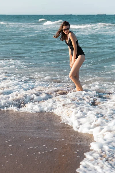 Femme heureuse en maillot de bain noir et des lunettes de soleil élégantes debout en mer — Photo de stock