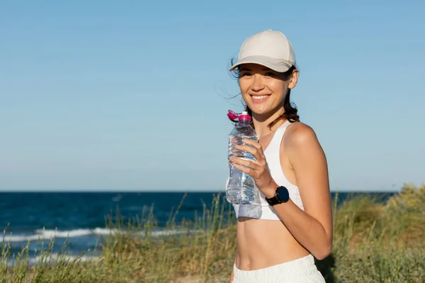 Happy and sportive woman in baseball cap holding bottle with water near ocean — Stock Photo