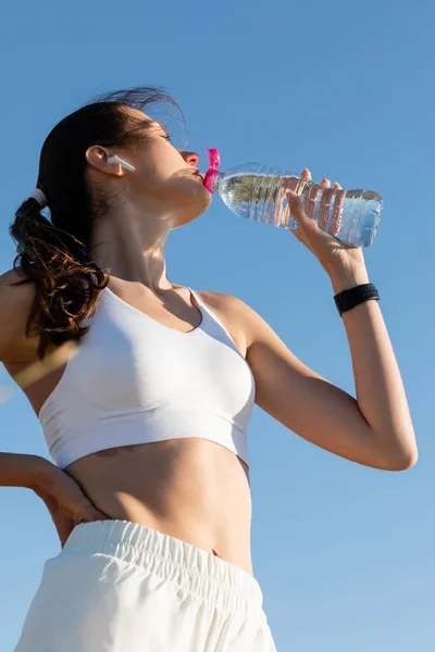 Low angle view of young woman in wireless earphone drinking water against blue sky — Stock Photo