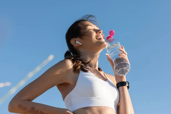 Low angle view of cheerful woman in wireless earphone holding bottle with water against blue sky — Stock Photo