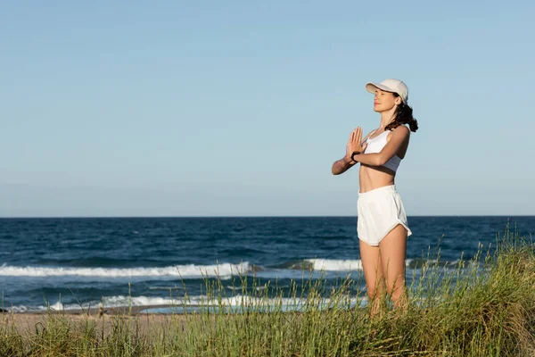 Young fit woman in shorts and sports bra meditating near blue sea on beach — Stock Photo