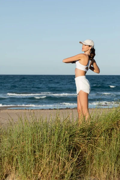 Mujer joven en forma en pantalones cortos y sujetador deportivo haciendo ejercicio cerca del mar azul en la playa - foto de stock