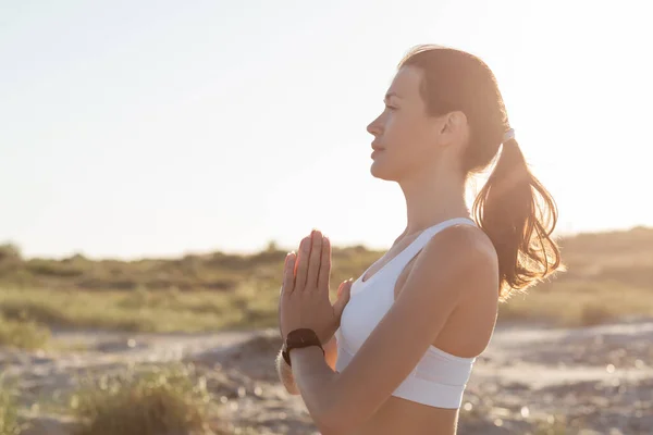 Side view of young fit woman in shorts and sports bra meditating with praying hands on beach in summer — Stock Photo