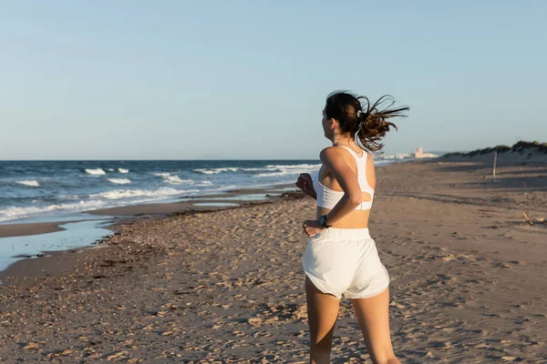 Young brunette woman in white sportswear jogging on sandy beach in summer — Stock Photo