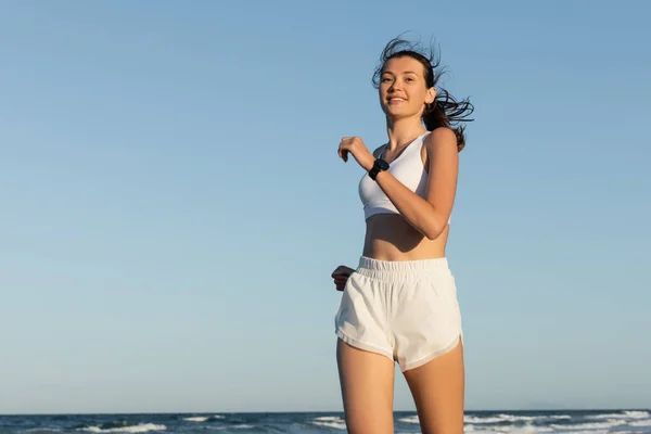 Vista de ángulo bajo de la joven mujer deportiva feliz trotando cerca del mar en verano - foto de stock